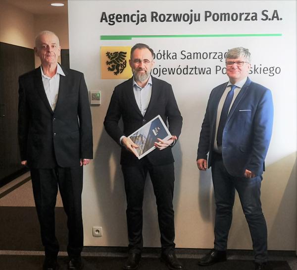 Prezesi zarządu ARP na wspólnej fotografii z grantobiorcą na tle ściany z logo Agencji Rozwoju Pomorza