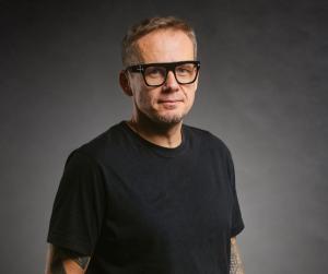 mężczyzna zfotografowany do pasa, na ciemnobrązowym tle, w okularach i w ciemnym t-shirtcie